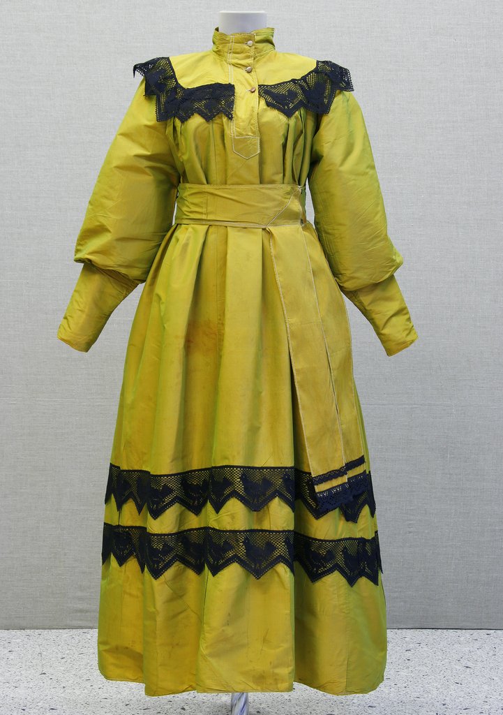 На манекене желто‑оливковое платье с высоким воротником-стойкой и пуговицами. Объëмные рукава-